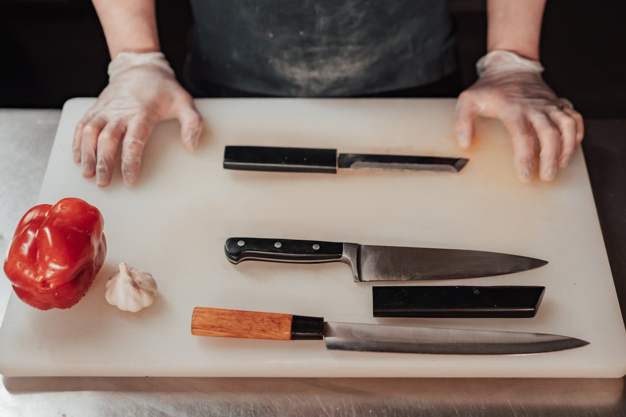 Gloved hands preparing to chop with knives on a cutting board next to vegetables in a kitchen scene.
